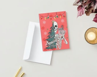 May include: A red Christmas card with a skeleton holding a mug and wearing a Christmas tree. The card has the text "Sorta Merry, Sorta Scary" and is next to a white envelope. A string of lights and snowflakes decorate the card.