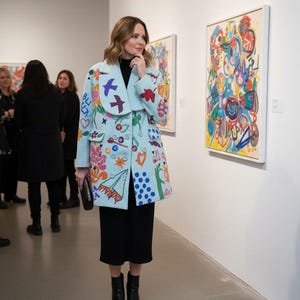May include: A light blue coat with colorful embroidered designs, including flowers, birds, and geometric shapes. The coat is worn over a black dress and paired with black boots. The setting appears to be an art gallery.