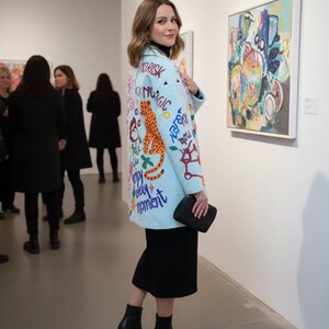 May include: A light blue coat with colorful graffiti-style text and a large orange leopard design. The coat is worn over a black outfit, and the person is holding a black clutch. The background shows an art gallery setting.