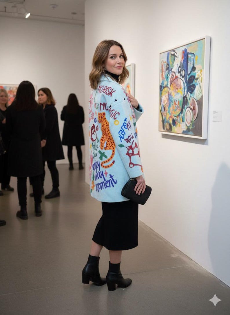 May include: A light blue coat with colorful graffiti-style text and a large orange leopard design. The coat is worn over a black outfit, and the person is holding a black clutch. The background shows an art gallery setting.