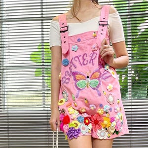 May include: Pink denim overalls with a butterfly and the word "Butterfly" embroidered on the front. The overalls are decorated with colorful crocheted flowers.