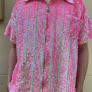 May include: A pink short-sleeved shirt with a zip closure and iridescent sequins. The shirt has a collar and a star-shaped zip pull.