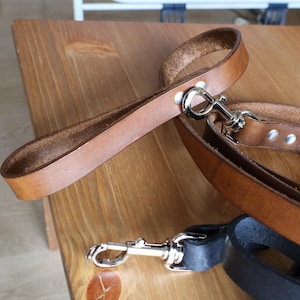 May include: A brown leather dog lead with a matching handle and a silver-coloured metal clasp. The lead is made of sturdy leather and is designed for durability. The handle is riveted to the lead.