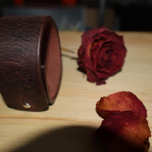 May include: A wide, dark brown leather cuff bracelet with a textured surface. A dried red rose and petals are next to the bracelet on a wooden surface. The bracelet is secured with a silver snap.
