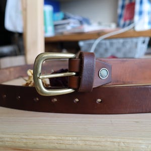 May include: A brown leather belt with a brass buckle. The belt has a classic design with a rectangular buckle and a single keeper loop. The belt is made of a sturdy leather material and has several holes for adjustment.