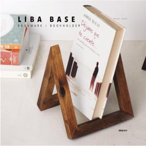 May include: A wooden book holder with a triangular design. The book holder is holding a book titled "Dejame que te cuente..." by Jorge Bucay. The book holder is made of dark brown wood and has a natural finish.
