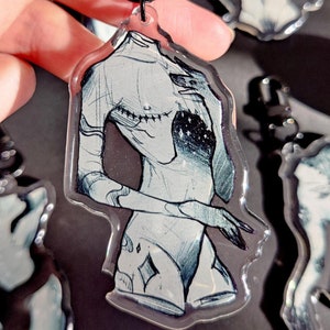 Puede incluir: Un llavero de acrílico transparente con un dibujo en blanco y negro de una figura sin cabeza con un diseño de galaxia en el pecho. La figura está de pie con los brazos cruzados y tiene un diamante en la cadera.