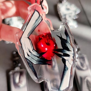 May include: A clear acrylic keychain with a black and white illustration of two hands holding a red apple. The keychain has a silver metal chain.