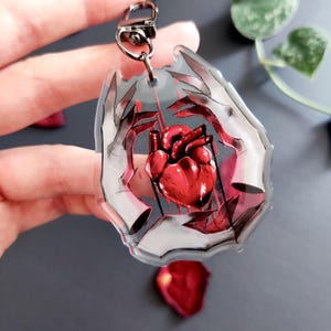 Puede incluir: Un llavero de acrílico transparente con un diseño de corazón anatómico rojo. El corazón está rodeado de manos estilizadas y detalles rojos. El llavero tiene un cierre plateado.