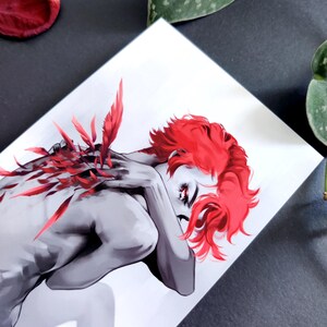 May include: An art print showing a grayscale figure with striking red hair and details. The figure is in a relaxed pose, with red feather-like elements. The artwork is set against a white background, with a dark surface.