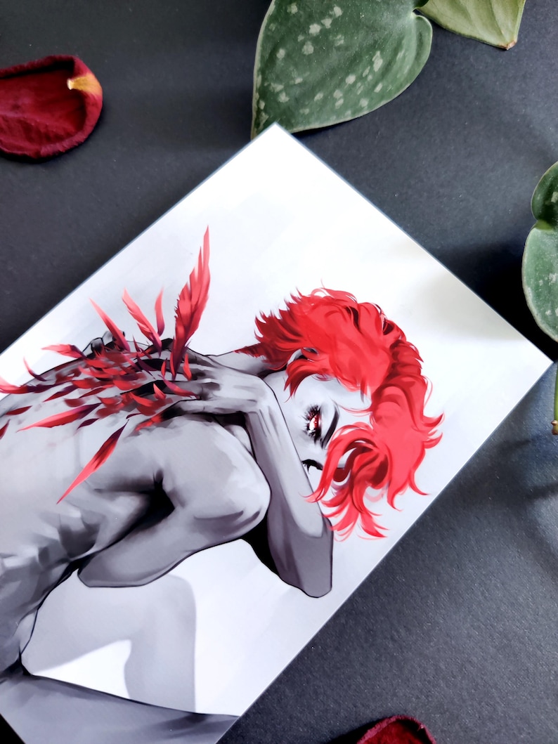 May include: An art print showing a grayscale figure with striking red hair and details. The figure is in a relaxed pose, with red feather-like elements. The artwork is set against a white background, with a dark surface.