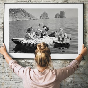 Puede incluir: Una fotografía en blanco y negro de tres mujeres en una balsa en el agua. Las mujeres llevan trajes de baño y sonríen. La balsa flota en el agua y hay montañas en el fondo.
