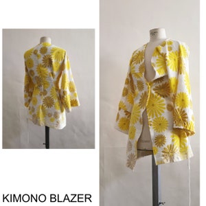 Puede incluir: Una chaqueta de kimono de estampado floral blanco y amarillo con mangas largas y cierre de corbata. La chaqueta está en un maniquí.