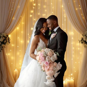 May include: A bride and groom stand close together, smiling, in front of a backdrop of sheer curtains and twinkling lights. The bride is wearing a white wedding dress and holding a bouquet of pink and white roses. The groom is wearing a black tuxedo.