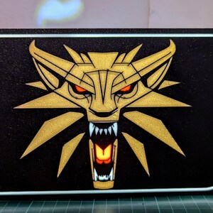 May include: A black and gold illuminated sign featuring a wolf's head graphic. The wolf's eyes are red and its mouth is open, revealing sharp teeth. The sign is lit from behind, creating a glowing effect.
