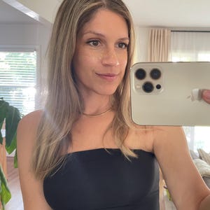 May include: A woman wearing a black tube top and a gold necklace, holding a smartphone. Her hair is long and blonde, and she has gold glitter on her face. The background includes a window, curtains, and a glimpse of a living room.