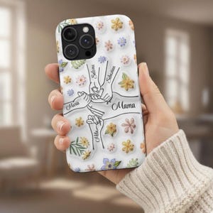 Puede incluir: Funda de teléfono blanca con diseño floral y las palabras "Mama", "Maria", "Salome", "Sebastian" y "Mila". La funda presenta un gráfico de manos entrelazadas. La funda tiene un recorte para la cámara.