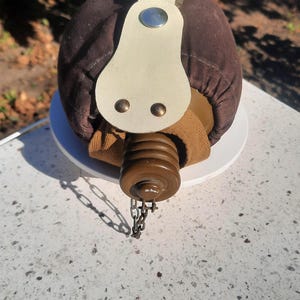 Puede incluir: Una cantimplora cubierta de tela marrón con una correa de cuero color crema y un botón de presión de metal plateado. La cantimplora tiene un tapón de rosca de plástico marrón unido a una pequeña cadena de metal y una correa de lona.