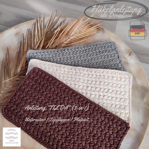 May include: Three crocheted square trivets in shades of grey, cream, and burgundy, arranged on a wooden tray with gold-coloured decorative leaves. The trivets have a textured, woven appearance. Text on the trivets reads "TILDA" and "Untersetzer / Topflappen / Platzset".