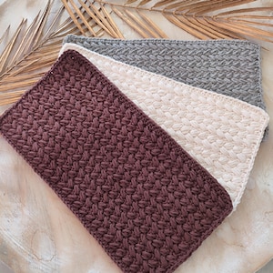 Könnte beinhalten: Drei rechteckige, handgehäkelte Waschlappen in den Farbtönen Burgunderrot, Creme und Grau. Jeder Lappen weist ein strukturiertes Webmuster auf. Die Waschlappen sind diagonal angeordnet und zeigen ihre unterschiedlichen Farben und Texturen.