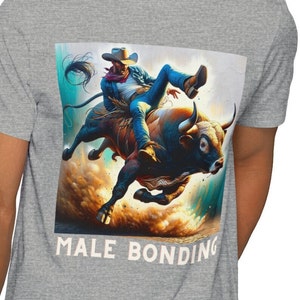 Puede incluir: Un vaquero con una camisa azul y pantalones vaqueros monta un toro marrón con la cara blanca. El toro está dando coces y levantando polvo. La imagen tiene el texto "MALE BONDING" en la parte inferior.