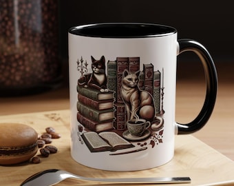 May include: A white ceramic mug with a black interior and handle. The mug features a detailed illustration of two cats, books, and a cup of coffee. The design is in shades of brown, black, and white, with a touch of gold.