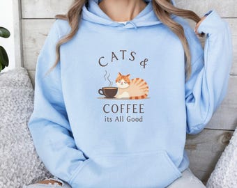 Puede incluir: Una sudadera con capucha azul claro con un gráfico de un gato bebiendo café y el texto "Cats & Coffee its All Good".