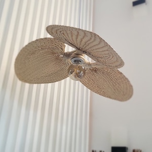 Puede incluir: Un ventilador de techo tejido con tres aspas, una bombilla y un fondo blanco. El ventilador está hecho de materiales naturales y tiene un aspecto rústico.