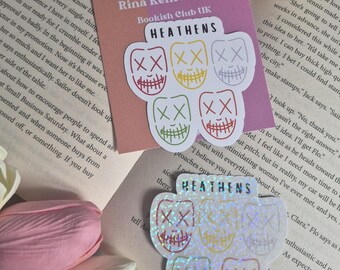May include: Two stickers featuring the text HEATHENS above five stylized faces with stitched mouths and X eyes. One sticker is on a square backing card labeled Rina Kent Sticker Bookish Club UK, and the other is a holographic version.