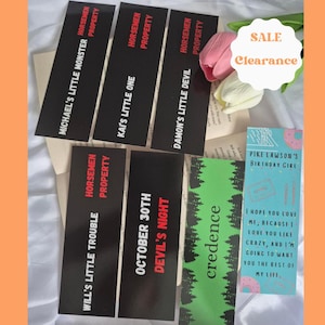 May include: A collection of rectangular bookmarks featuring black backgrounds with red and white text including phrases like Michael's Little Monster, Kai's Little One, Damon's Little Devil, Will's Little Trouble, October 30th Devil's Night, Credence, and Pike Lawson's Birthday Girl.