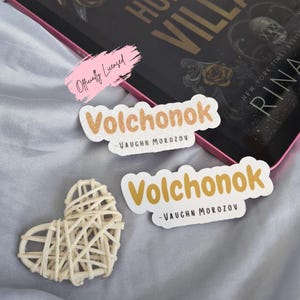May include: Two stickers with the word "Volchonok" in a playful font, with "-VAUGHN MOROZOV" below. A woven heart and a book titled "HUNT THE VILLAIN" are also visible. The stickers have a light brown and white color scheme.