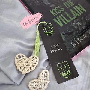 May include: A black bookmark with a neon green face design and the text "Little Monster" is next to a book titled "Kiss the Villain". Two woven heart-shaped decorations and a pink "Officially Licensed" sticker are also visible.