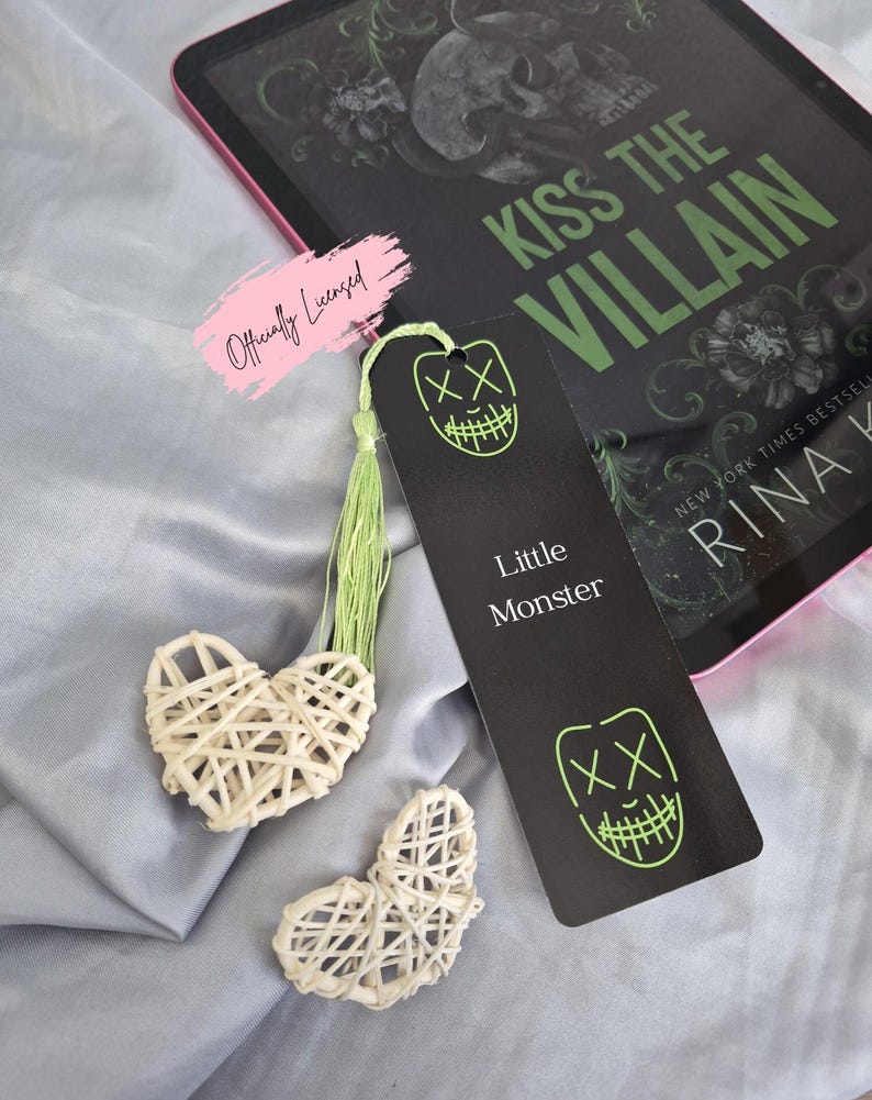 May include: A black bookmark with a neon green face design and the text "Little Monster" is next to a book titled "Kiss the Villain". Two woven heart-shaped decorations and a pink "Officially Licensed" sticker are also visible.