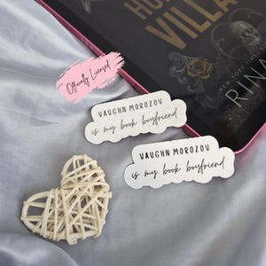 May include: A book and stickers on a gray satin surface. The book is titled "Hunt the Villain" and has gold lettering. Two stickers read "Vaughn Morozov is my book boyfriend." A woven heart is also present.