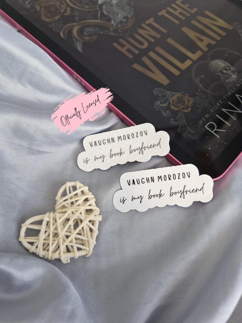 May include: A book and stickers on a gray satin surface. The book is titled "Hunt the Villain" and has gold lettering. Two stickers read "Vaughn Morozov is my book boyfriend." A woven heart is also present.