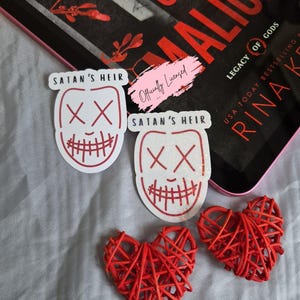 May include: The image features two white stickers with a red outline of a mask design and the text "SATAN'S HEIR". Two red woven hearts and a book titled "GODS OF MALICE" are also visible. The book cover has red text.