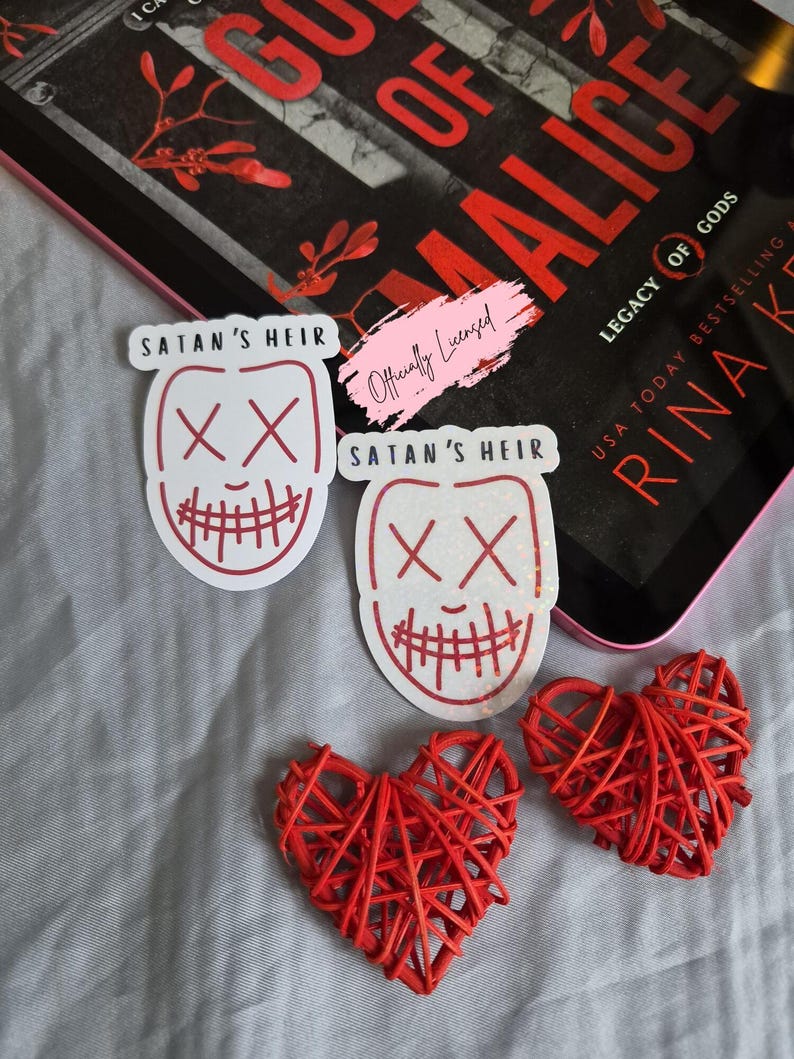 May include: The image features two white stickers with a red outline of a mask design and the text "SATAN'S HEIR". Two red woven hearts and a book titled "GODS OF MALICE" are also visible. The book cover has red text.