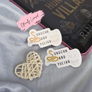 May include: A collection of stickers and a woven heart on a light blue surface. The stickers feature text and graphics, including a snake, a mask, and the words "VAUGHN AND YULIAN." A book with the title "HUNT THE VILLAIN" is also visible.