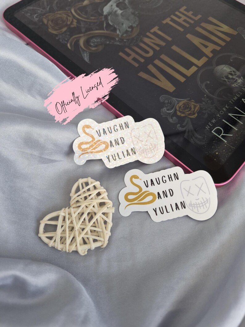 May include: A collection of stickers and a woven heart on a light blue surface. The stickers feature text and graphics, including a snake, a mask, and the words "VAUGHN AND YULIAN." A book with the title "HUNT THE VILLAIN" is also visible.