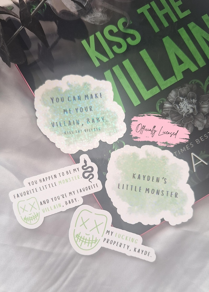 May include: A collection of white stickers with green and black text, featuring phrases like "You can make me your villain, baby" and "Kayden's little monster." The stickers are on a book with the title "Kiss the Villain."