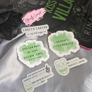 May include: A collection of five die-cut vinyl stickers featuring green watercolor accents and black text. Phrases include Gareth Carson is my book boyfriend, You can make me your villain, baby, Kayden's little monster, and My fucking property, Kayde.