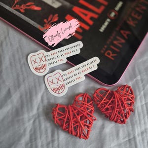 May include: An image featuring a book titled "Gods of Alice" by Rina Kent, accompanied by two stickers with a graphic design and the text "I'LL MAKE SURE YOU ALWAYS CHOOSE ME AS MUCH AS I CHOOSE YOU." Two red woven hearts are also present.