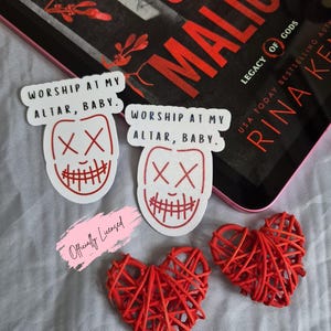 May include: A collection of items including stickers, and heart-shaped decorations. The stickers feature a red and white design with the text "WORSHIP AT MY ALTAR, BABY." Two red woven hearts are also present, along with a book.
