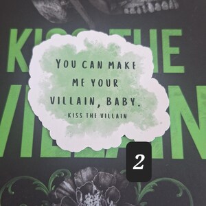 May include: A book cover with a skull and floral design. A sticker with the text "YOU CAN MAKE ME YOUR VILLAIN, BABY. - KISS THE VILLAIN" is placed on the cover. The background is black and green.