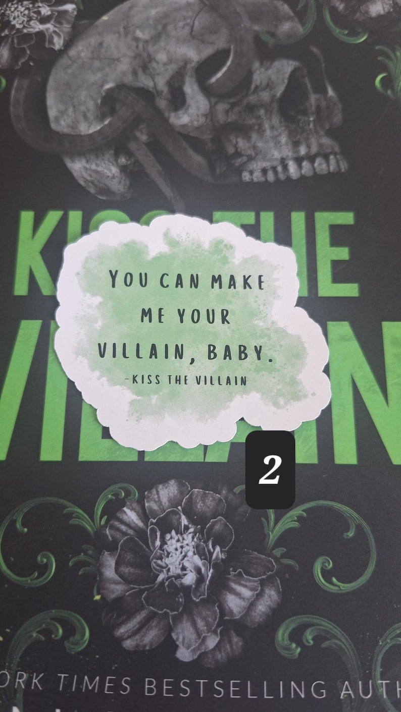 May include: A book cover with a skull and floral design. A sticker with the text "YOU CAN MAKE ME YOUR VILLAIN, BABY. - KISS THE VILLAIN" is placed on the cover. The background is black and green.