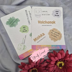May include: Two white rectangular cards with text and graphics, including "Volchonok" and "Mishka." One card has "Gareth Carson" and "Kayden's Little Monster." The cards are on a gray surface with red flowers.