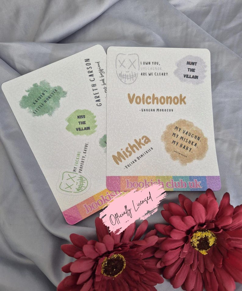 May include: Two white rectangular cards with text and graphics, including "Volchonok" and "Mishka." One card has "Gareth Carson" and "Kayden's Little Monster." The cards are on a gray surface with red flowers.