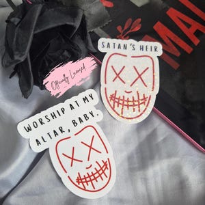 May include: Two white stickers with red outlines and text. One sticker reads "WORSHIP AT MY ALTAR, BABY." and features a face with an "XX" eye design. The other sticker says "SATAN'S HEIR" and has a similar face design.