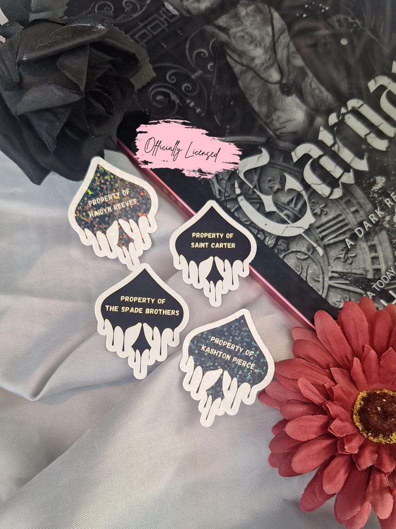 Puede incluir: Cuatro pegatinas en blanco y negro con un dise&ntilde;o de goteo y texto dorado que dice "Property of Haidyn Reeves", "Property of Saint Carter", "Property of The Spade Brothers" y "Property of Kashton Pierce". Las pegatinas est&aacute;n sobre una superficie gris claro.