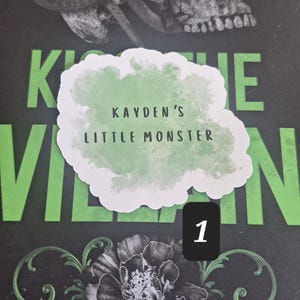 May include: A book cover with a skull and snake design, with the text "KAYDEN'S LITTLE MONSTER" in a cloud-shaped graphic. The background is black with green accents and floral illustrations.